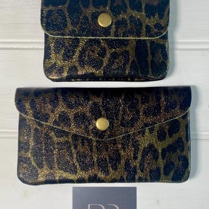 May include: Two small, black and gold leopard print wallets with a snap closure. The wallets are rectangular with rounded edges. The top wallet is smaller than the bottom wallet. The background is a white wooden surface.