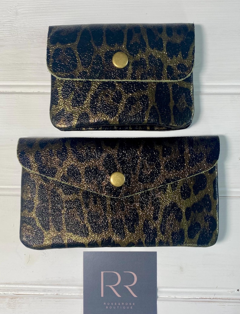 May include: Two small, black and gold leopard print wallets with a snap closure. The wallets are rectangular with rounded edges. The top wallet is smaller than the bottom wallet. The background is a white wooden surface.