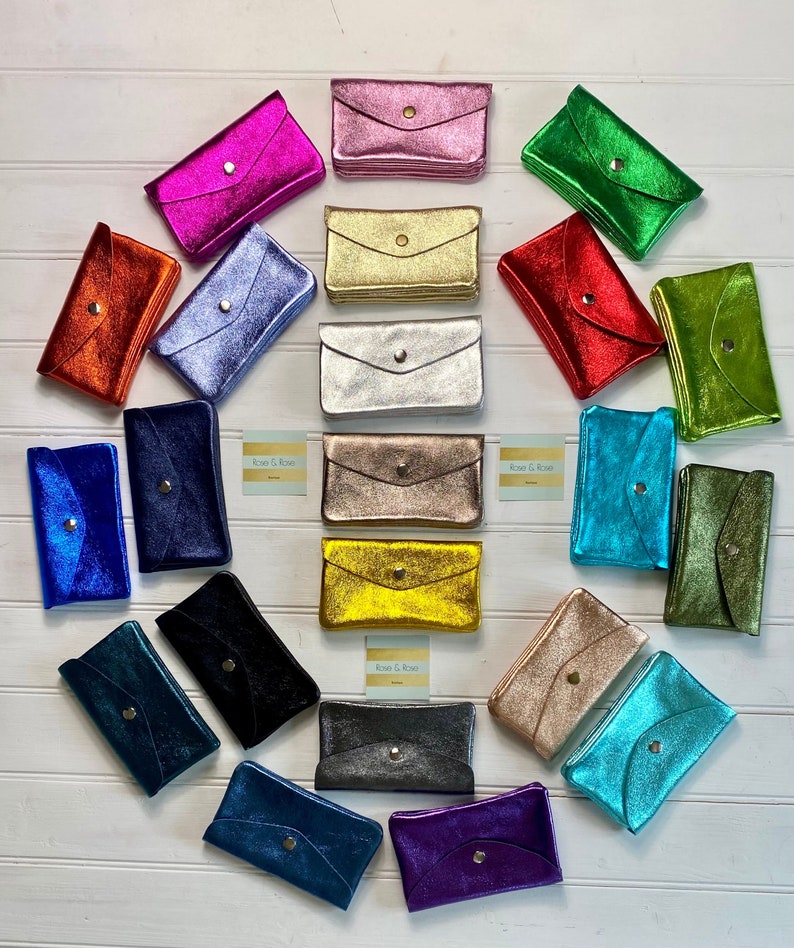 May include: A selection of metallic leather envelope wallets in shades like pink, gold, silver, red, and blue. Each wallet has a snap closure and a textured finish. The wallets are displayed on a white wooden surface.