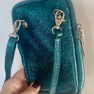May include: A teal-colored, metallic crossbody bag with a textured finish. The bag features a silver zipper, silver clasps, and a matching teal strap. The bag is designed to hold a phone and other small items.