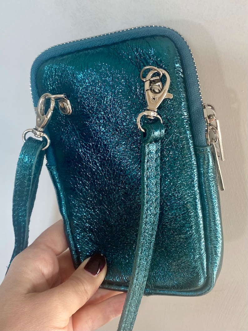 May include: A teal-colored, metallic crossbody bag with a textured finish. The bag features a silver zipper, silver clasps, and a matching teal strap. The bag is designed to hold a phone and other small items.