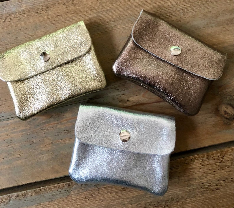 May include: Three small, metallic leather coin purses in gold, bronze, and silver. Each purse has a flap closure secured by a silver snap. The purses are arranged on a wooden surface, showcasing their textured appearance.