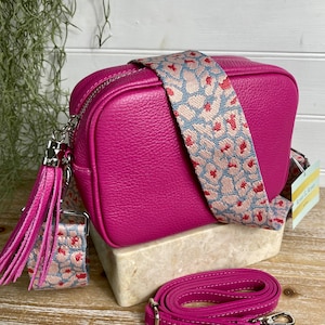 Könnte beinhalten: Fuchsiafarbene Umhängetasche mit einem gemusterten Riemen mit einem floralen Design in Rosa und Blau. Die Tasche hat eine passende Quaste und einen zusätzlichen verstellbaren Riemen. Die Tasche wird auf einem Steinblock präsentiert.