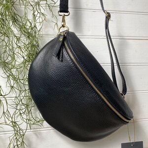 May include: Black leather crossbody bag with a curved design and a gold zipper. The bag has a long, adjustable strap and a small tag attached. The bag is hanging against a white wooden background with green plants.