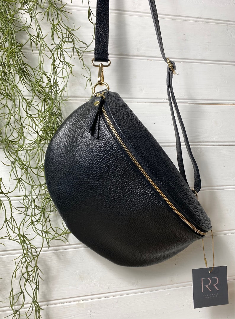 May include: Black leather crossbody bag with a curved design and a gold zipper. The bag has a long, adjustable strap and a small tag attached. The bag is hanging against a white wooden background with green plants.