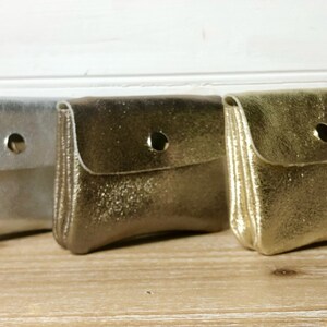 May include: Three small, metallic coin purses in silver, bronze, and gold. Each purse has a flap closure secured by a single snap. The purses are displayed on a light-coloured wooden surface.