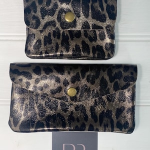 May include: Two small, rectangular leopard print pouches with a gold metallic sheen. Each pouch has a flap closure secured by a gold-coloured snap. The pouches are displayed on a white wooden surface.