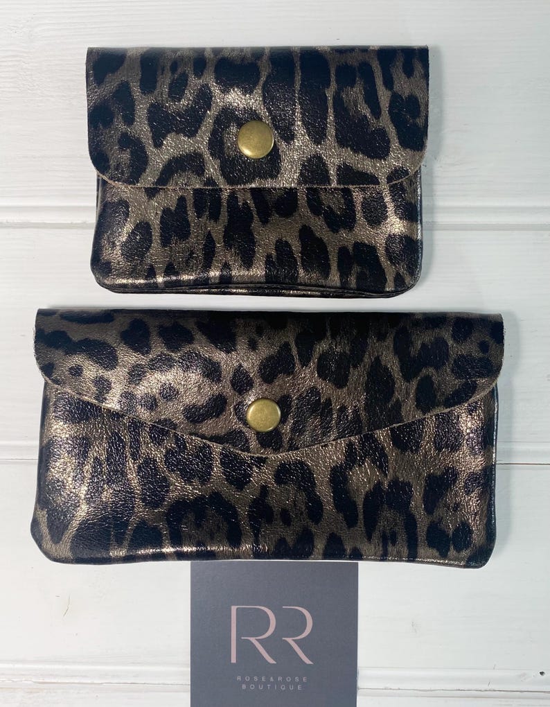 May include: Two small, rectangular leopard print pouches with a gold metallic sheen. Each pouch has a flap closure secured by a gold-coloured snap. The pouches are displayed on a white wooden surface.