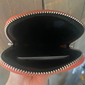 May include: Open orange zippered pouch with a silver zipper. The interior is black, and a smartphone is visible inside. The pouch is made of a textured material.