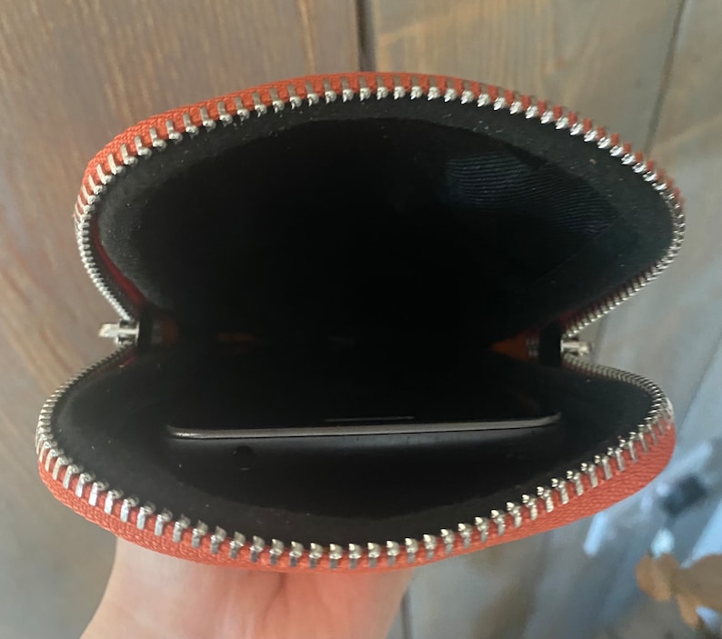 May include: Open orange zippered pouch with a silver zipper. The interior is black, and a smartphone is visible inside. The pouch is made of a textured material.