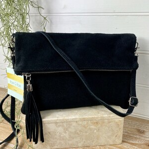 May include: Black suede crossbody bag with a fold-over flap and a long, adjustable strap. The bag features a silver zipper and a decorative tassel. The bag is sitting on a marble-like box.