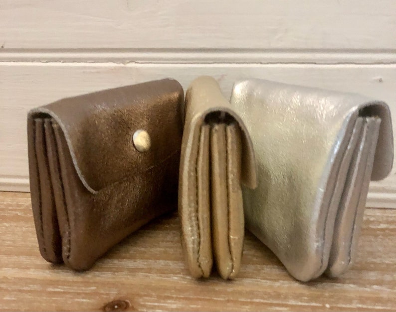 May include: Three leather wallets in metallic shades of bronze, gold, and silver. The bronze wallet has a snap closure. The wallets are folded and have multiple compartments, suggesting they are designed for cards and cash.