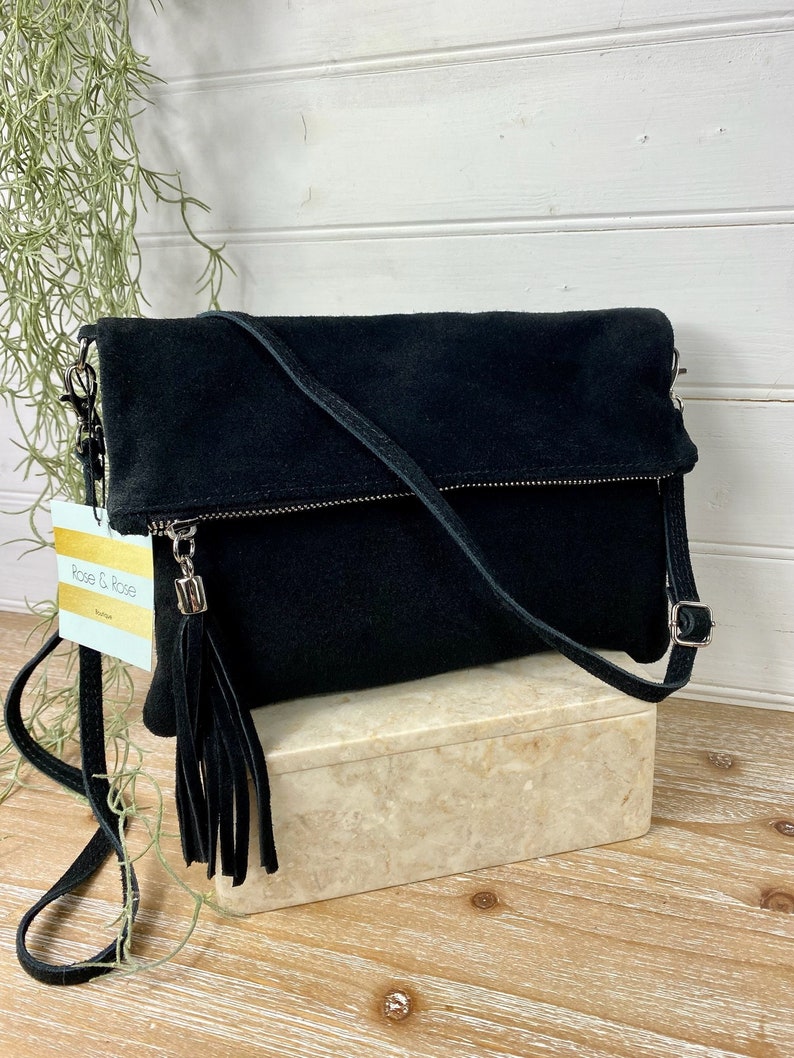 May include: Black suede crossbody bag with a fold-over flap and a long, adjustable strap. The bag features a silver zipper and a matching tassel. The tag reads "Rose & Rose Boutique".