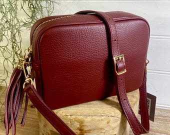 May include: A burgundy leather crossbody bag with a rectangular shape. The bag features a gold zipper, a matching tassel, and a detachable strap. The bag is sitting on a small wooden stool.