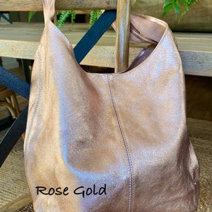 May include: Rose gold leather shoulder bag with a single strap. The bag is displayed on a wooden chair. The text "Rose Gold" is written on the bag.