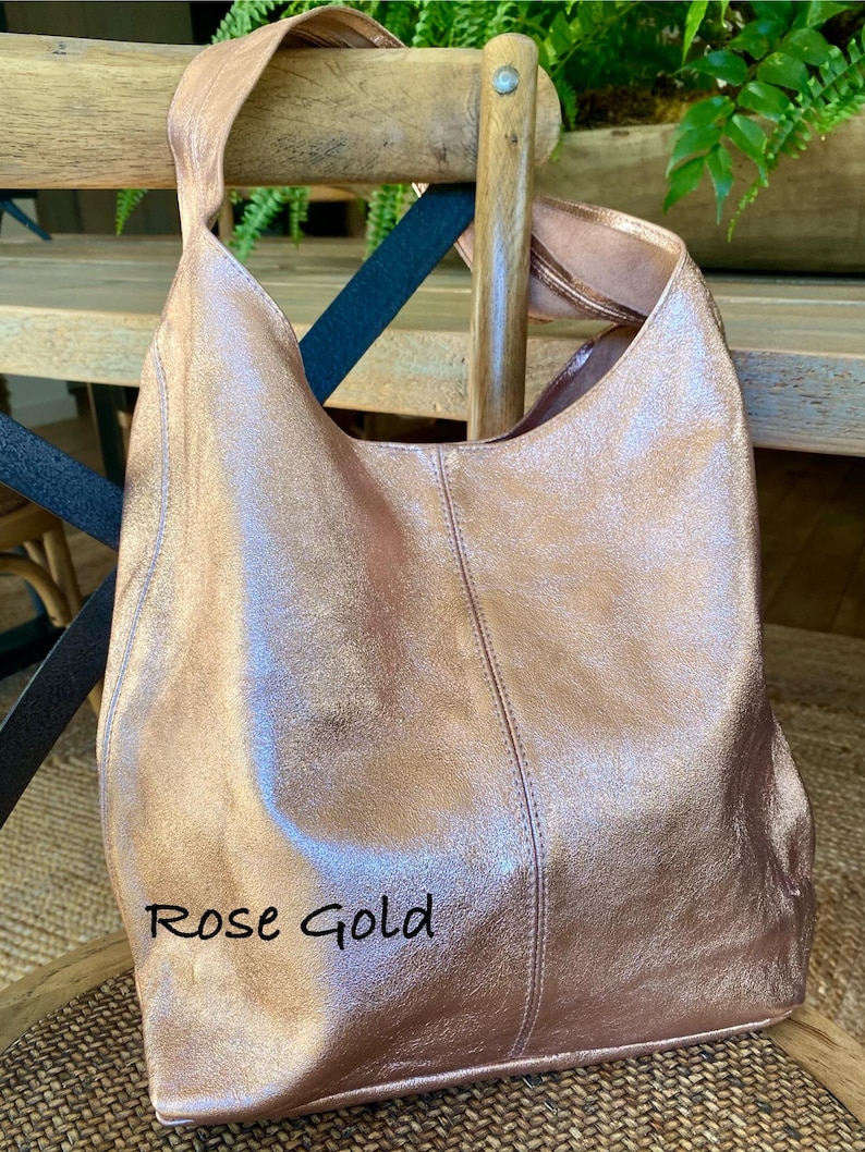 May include: Rose gold leather shoulder bag with a single strap. The bag is displayed on a wooden chair. The text "Rose Gold" is written on the bag.