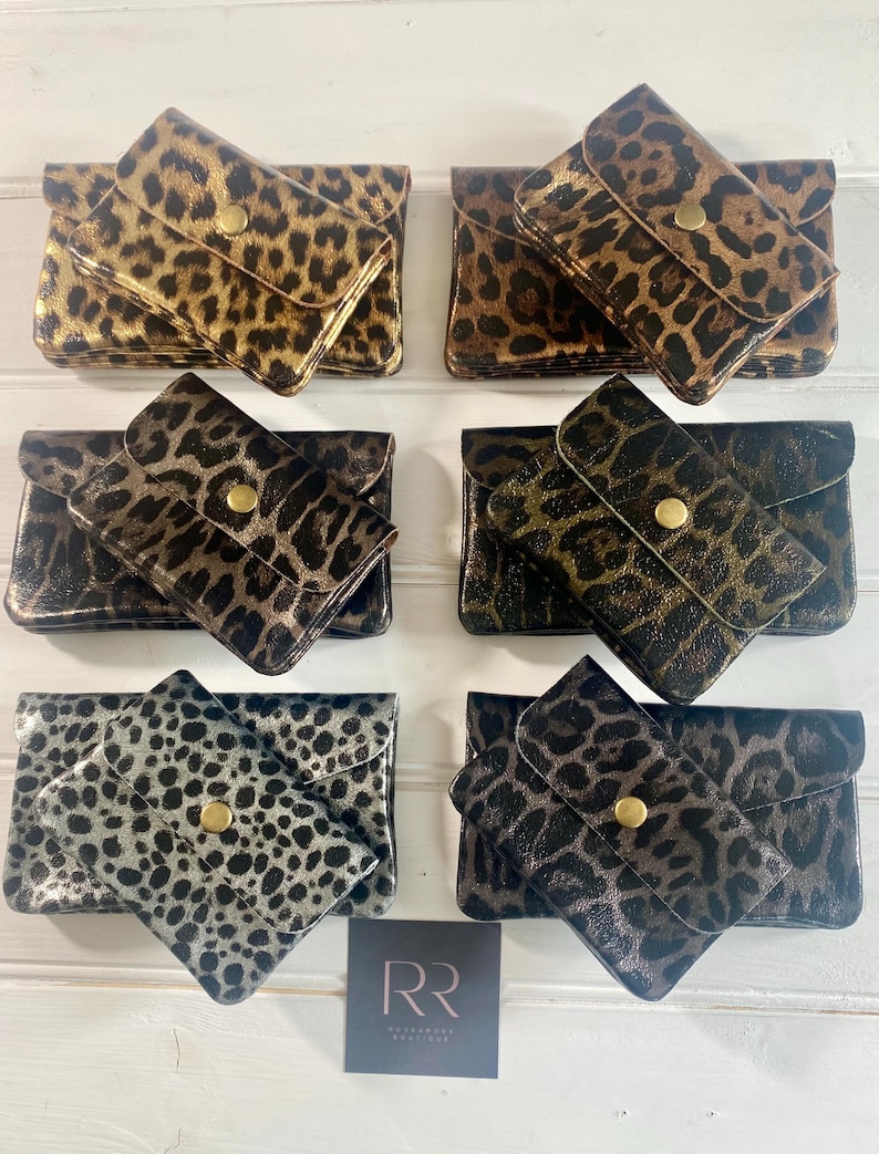 May include: Six small, rectangular leopard print wallets in shades of gold, brown, and silver. Each wallet has a flap closure with a gold button. The wallets are arranged on a white wooden surface. A small card with the letters "RR" is in the bottom right corner.