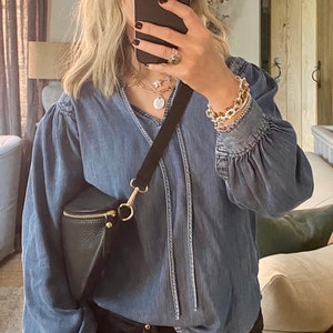 May include: A denim blue blouse with long sleeves and a V-neck, paired with black jeans. A black crossbody bag with a gold zipper and hardware is worn across the body. The person is wearing gold jewelry and a watch.