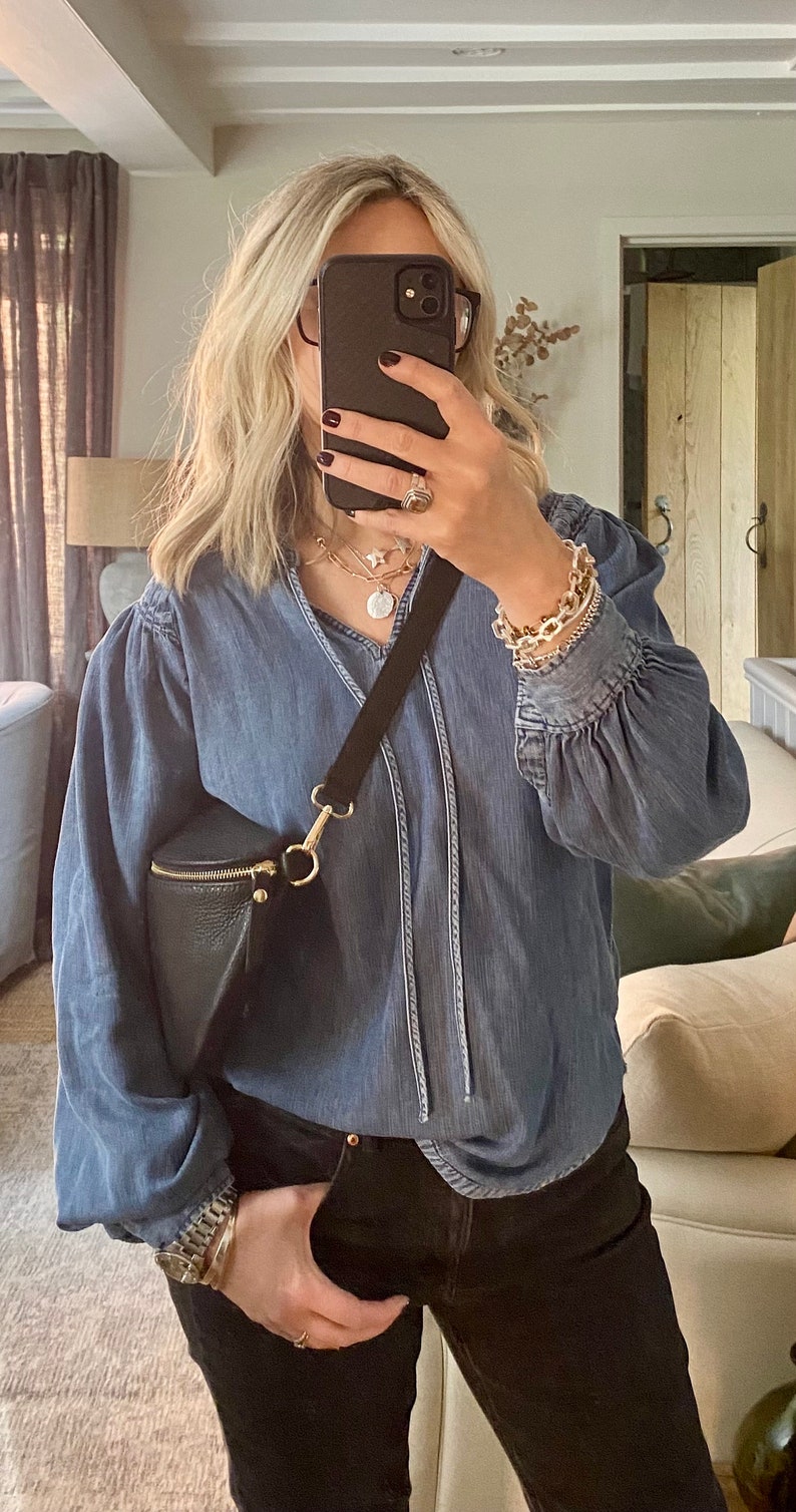 May include: A denim blue blouse with long sleeves and a V-neck, paired with black jeans. A black crossbody bag with a gold zipper and hardware is worn across the body. The person is wearing gold jewelry and a watch.