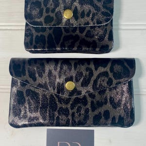 May include: Two black and grey leopard print wallets with gold-coloured snap closures. The wallets are different sizes, with a smaller one on top and a larger one below. A small card with the text "RR ROSE & ROSE BOUTIQUE" is at the bottom.