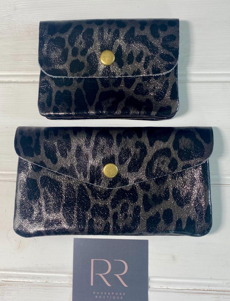 May include: Two black and grey leopard print wallets with gold-coloured snap closures. The wallets are different sizes, with a smaller one on top and a larger one below. A small card with the text "RR ROSE & ROSE BOUTIQUE" is at the bottom.