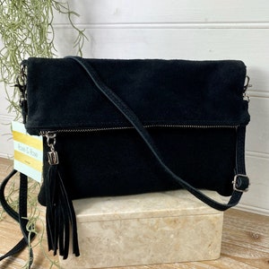 May include: Black suede crossbody bag with a fold-over flap and a long, adjustable strap. The bag features a silver zipper and a decorative tassel. The bag is sitting on a marble-like box.