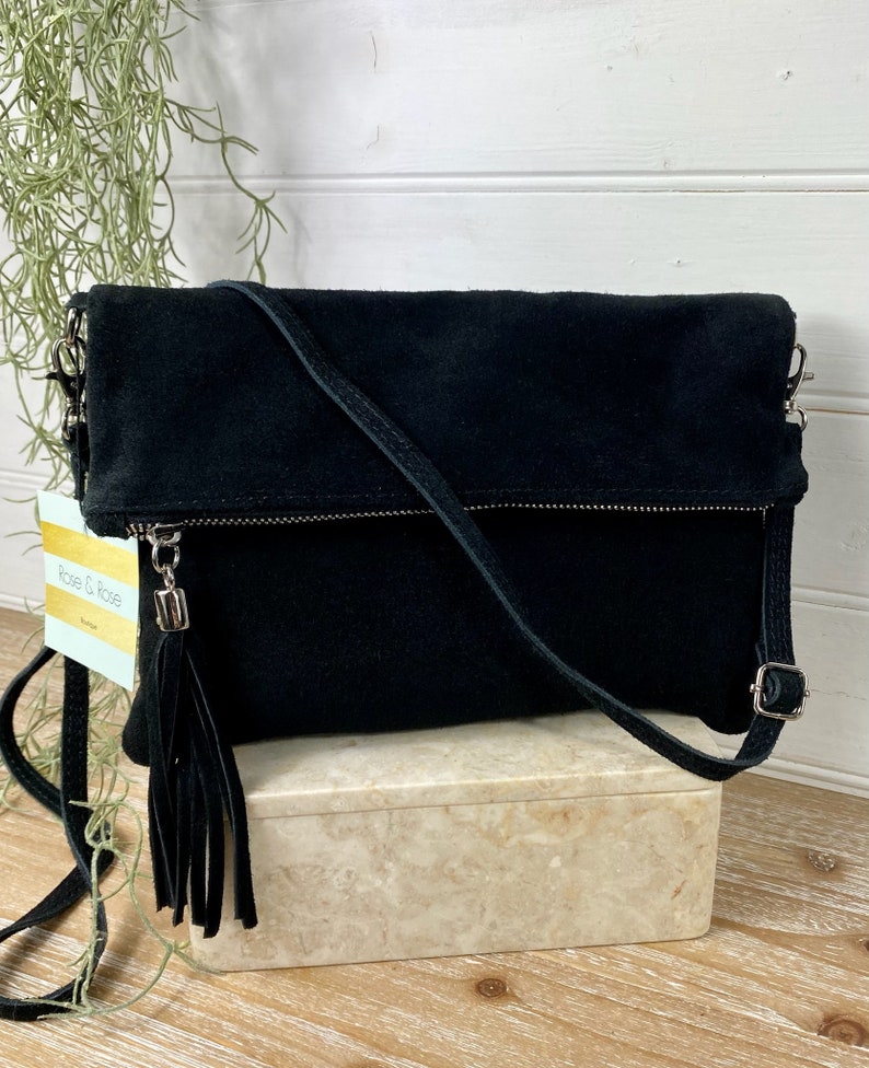May include: Black suede crossbody bag with a fold-over flap and a long, adjustable strap. The bag features a silver zipper and a decorative tassel. The bag is sitting on a marble-like box.