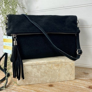 May include: Black suede crossbody bag with a fold-over flap, silver-tone hardware, and a long, adjustable strap. The bag features a zipper closure and a decorative tassel. The tag reads "Rose & Rose".