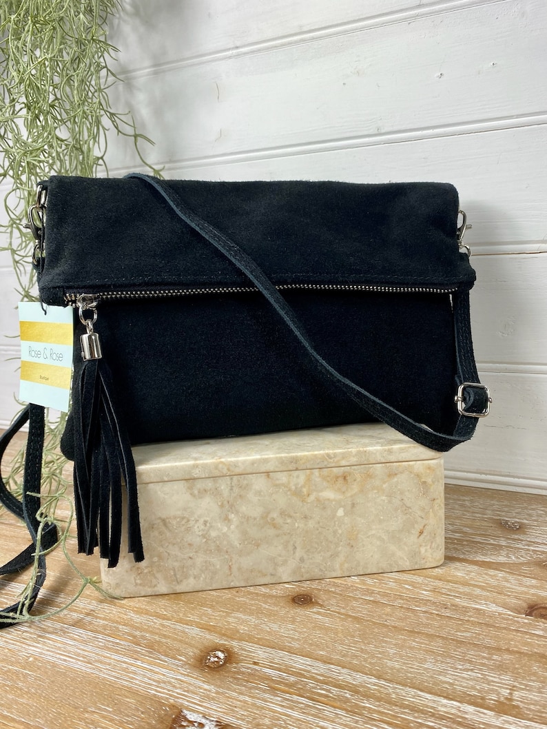 May include: Black suede crossbody bag with a fold-over flap, silver-tone hardware, and a long, adjustable strap. The bag features a zipper closure and a decorative tassel. The tag reads "Rose & Rose".