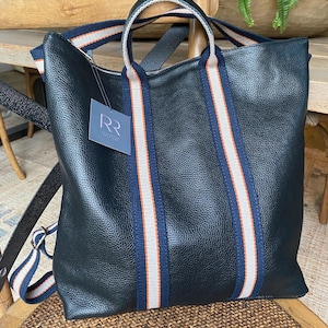 May include: Black leather tote bag with striped straps in navy, orange, and cream. The bag has a zipper closure and a tag attached. The bag can be carried by the top handles or worn as a backpack.