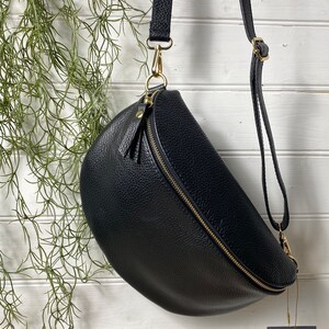 May include: Black leather crossbody bag with a gold zipper and hardware. The bag has a curved shape and a long adjustable strap. A small tag with the letters "RR" hangs from the bag.