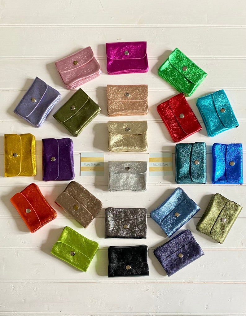May include: A selection of small, shimmering wallets in a variety of colours, such as pink, green, gold, and blue. Each wallet features a flap closure with a single button. The wallets are displayed on a white wooden surface.