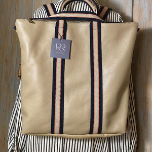 Könnte beinhalten: Ein beigefarbener Lederrucksack mit Reißverschluss oben, zwei Tragegriffen und zwei vertikalen Riemen in Marineblau mit einem dünnen orangefarbenen Mittelstreifen. Ein quadratisches Etikett mit den Buchstaben RR ist an der Vorderseite befestigt.