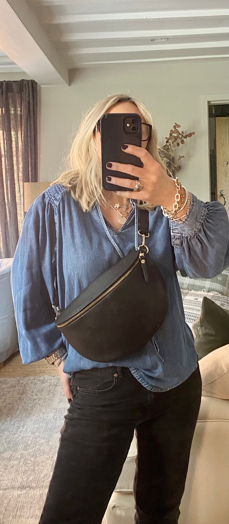 May include: Black leather crescent-shaped fanny pack with a gold zipper and hardware. The bag is worn across the body. The person is wearing a denim shirt and black jeans.