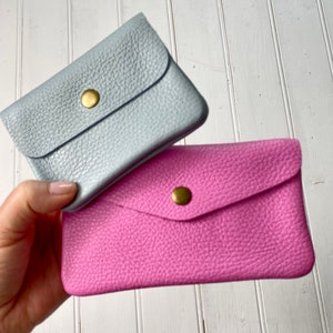 May include: Two small leather wallets, one light blue and one pink, are stacked. Each wallet has a flap closure secured by a gold-colored snap. The leather has a textured grain.