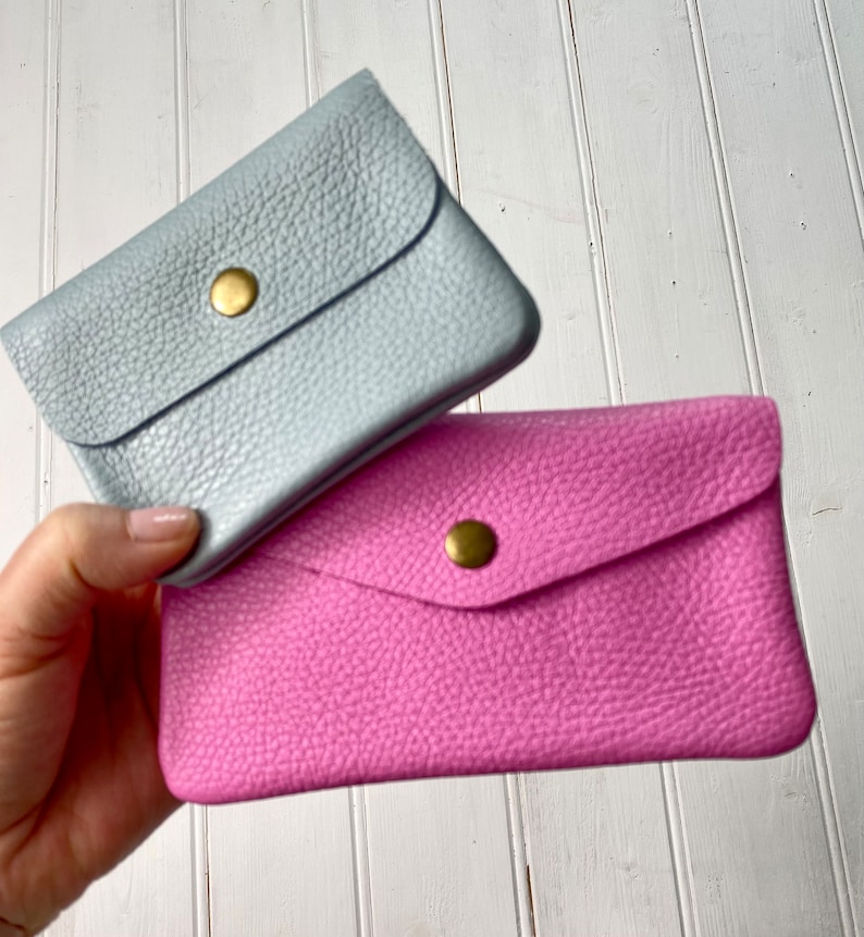 May include: Two small leather wallets, one light blue and one pink, are stacked. Each wallet has a flap closure secured by a gold-colored snap. The leather has a textured grain.