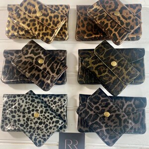 May include: Six small, rectangular leopard print wallets in shades of gold, brown, and silver. Each wallet has a flap closure with a gold button. The wallets are arranged on a white wooden surface. A small card with the letters "RR" is in the bottom right corner.