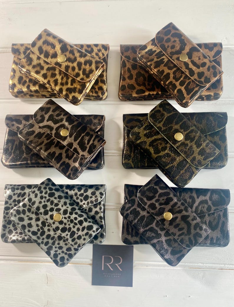 May include: Six small, rectangular leopard print wallets in shades of gold, brown, and silver. Each wallet has a flap closure with a gold button. The wallets are arranged on a white wooden surface. A small card with the letters "RR" is in the bottom right corner.