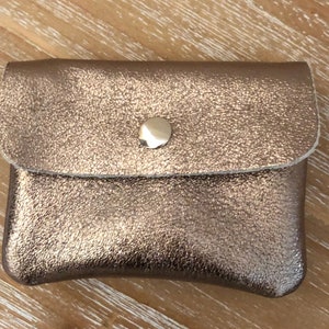 May include: A small, metallic bronze leather coin purse with a snap closure. The purse has a textured surface and a rounded flap. The snap is a shiny, gold-coloured metal. The purse is rectangular with rounded edges.