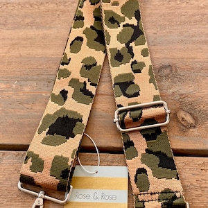 May include: A patterned bag strap with a tan background and green and black spots. The strap has silver-colored metal hardware and a tag that reads "Rose & Rose Boutique."