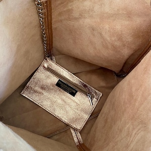 May include: A metallic rose gold tote bag with a matching interior. A small, rectangular zippered pouch is attached inside with a chain. The bag has a magnetic snap closure. The words "GENUINE LEATHER" are printed on a black label on the pouch.