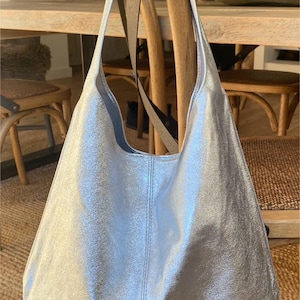 May include: A silver tote bag with a metallic finish. The bag has a shoulder strap and the word "silver" is written on the front. The bag is hanging on a wooden chair.