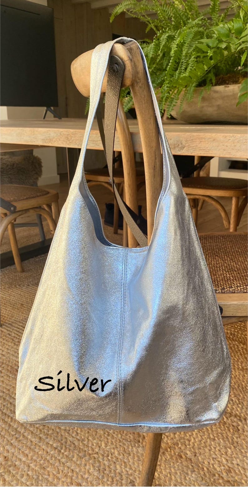May include: A silver tote bag with a metallic finish. The bag has a shoulder strap and the word "silver" is written on the front. The bag is hanging on a wooden chair.