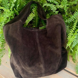 May include: A dark brown suede tote bag with a single handle. The bag has a soft, slouchy design and visible stitching details. The bag is set against a backdrop of green foliage.