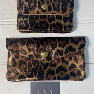 May include: Two leopard print clutch purses with gold-coloured hardware. The purses are in different sizes, with a small square purse on top and a larger rectangular purse below. The purses have a gold-coloured button closure.