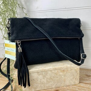 May include: Black suede crossbody bag with a silver zipper and tassel. The bag has a long, adjustable strap and a fold-over flap. The tag reads "Rose & Rose Boutique". The bag is displayed on a decorative box.