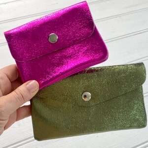 May include: Two small, metallic wallets. One is a vibrant magenta, and the other is olive green. Each wallet has a flap closure secured by a silver press stud. The wallets have a textured, crinkled appearance.
