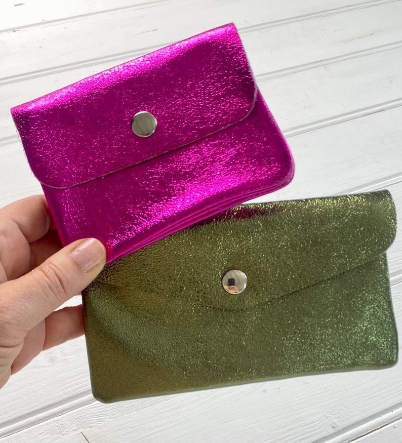 May include: Two small, metallic wallets. One is a vibrant magenta, and the other is olive green. Each wallet has a flap closure secured by a silver press stud. The wallets have a textured, crinkled appearance.