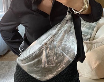 May include: A silver metallic bum bag with two zipped compartments and a long adjustable strap. The bag has a textured, crinkled appearance and is worn across the body. The bag is made of a shiny, reflective material.
