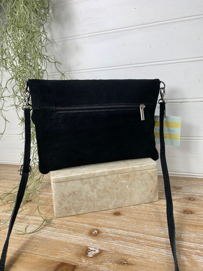 May include: A black suede crossbody bag with a silver chain strap. The bag has a zipper closure and a small tag that says "Rose & Rose". The bag is sitting on a marble-like box.